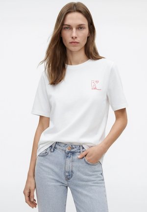 Young woman wearing a white t-shirt with a small red cat and heart design on the chest, paired with light blue jeans.