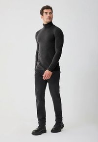 Schwarzes Rollkragenhemd mit figurbetontem Design und langen Ärmeln, kombiniert mit dunkler Hose und schwarzen Stiefeln, vor einem schlichten Hintergrund.