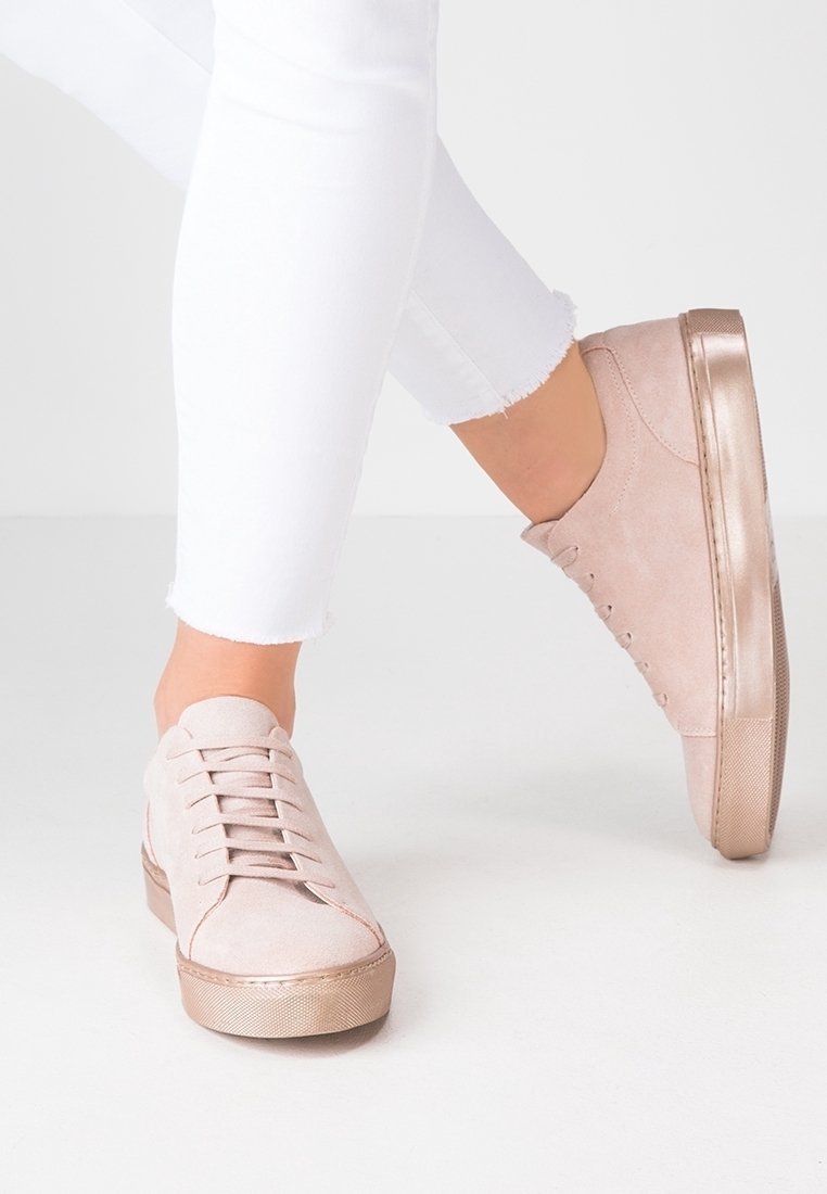 Personne portant un pantalon blanc à ourlet effiloché et des baskets en daim rose clair avec des semelles métalliques en or rose, se tenant sur un sol blanc.