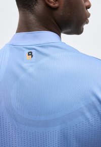 Κοντινή εικόνα στον άνω μέρος της πλάτης ενός ατόμου που φοράει ανοιχτό μπλε αθλητικό μπλουζάκι με υφή και μικρό λογότυπο κοντά στο λαιμό.