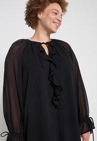 Černá halenka s dlouhými rukávy vyrobená z lehké tkaniny, s volánkovým předním dílem, zavazovacím výstřihem a nařasenými manžetami s elastickým detailem.