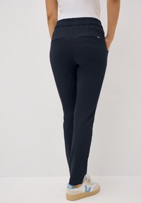 Marineblauwe taps toelopende broek met een elastische tailleband, twee achterzakken en een gladde textuur, gecombineerd met lichtgekleurde sneakers.