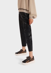 Persona che indossa pantaloni in pelle nera, felpa oversize beige e sneakers con piattaforma a stampa leopardata, in piedi su uno sfondo bianco.