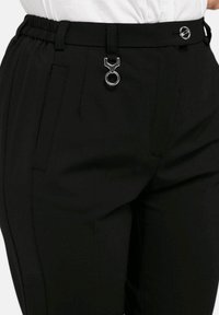 Zwarte broek met elastische tailleband, voorzien van een zilveren ringaccent aan de voorkant en zijzakken. Gladde textuur, op maat gemaakt ontwerp.