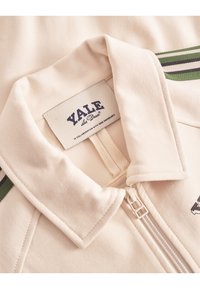 Kremfarget zip-up jakke med krage, med grønne og hvite striper, og en "Yale"-lapp ved kragen. Glatt stofftekstur.