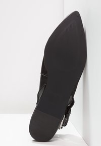 Semelle de botte noire à bout pointu avec talon texturé et grip en forme de boule, posée contre un mur d'angle blanc et un sol.