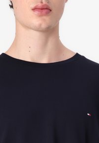 Nærbillede af en persons underansigt og hals iført en mørkeblå skjorte med et lille rødt, hvidt og blåt logo på brystet.