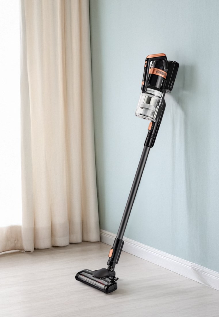 Kabelloser Staubsauger mit schwarzem Gehäuse, transparentem Staubbehälter und orangefarbenen Akzenten, der an einer hellblauen Wand mit Vorhängen lehnt.