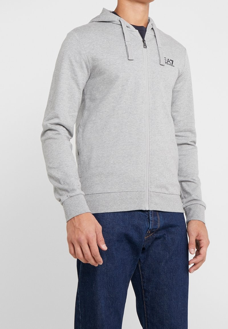 Sudadera con cremallera de color gris claro, hecha de una suave mezcla de algodón. Tiene capucha, bolsillos delanteros y puños acanalados. Pequeño logo en el pecho, ajuste casual.