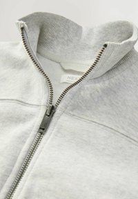 Veste zippée grise avec un col haut. Dotée d'une fermeture éclair métallique et d'une texture en côtes au niveau du col. Le tissu est lisse et légèrement texturé.