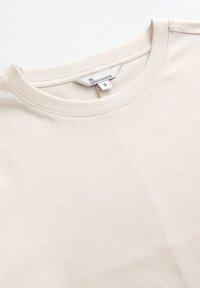 T-shirt in cotone beige con scollo rotondo, maniche corte e una texture morbida. Include un'etichetta con le informazioni sulla taglia e la collezione.