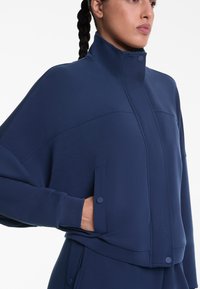 Navyblaue kurz geschnittene Jacke aus weichem Stoff mit einem hohen Kragen, Fronttaschen und dezenten Nähdetails. Glatte Textur und modernes Design.