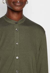 Olijfgroene shirt met hoge kraag en vijf knoopsluiting, met ronde knopen en een zachte textuur.