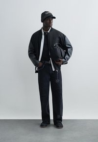 Zwarte leren bomberjack over een zwart T-shirt, gecombineerd met donkere jeans met versieringen. Accessoires omvatten een geruite pet en tas.