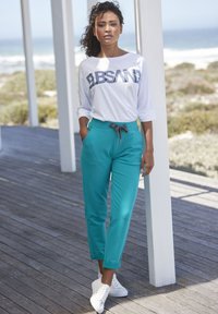 Weißes Langarmhemd mit grafischem Druck, kombiniert mit hellblauen, lässig sitzenden Hosen. Sneakers runden den entspannten Look ab. Holzdeck im Hintergrund.