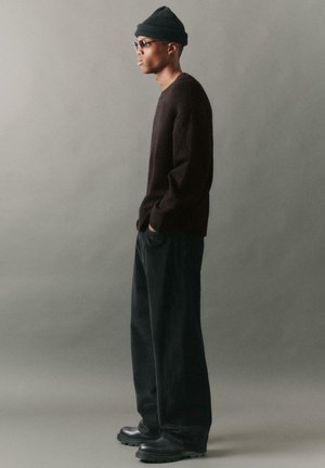 Pullover in pile scuro, pantaloni in corduroy nero a coste e stivaletti neri alla caviglia. Gli accessori includono un berretto lavorato a maglia e occhiali. Vista laterale.