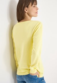 Femme aux cheveux bruns portant un pull à manches longues jaune clair et un jean bleu, debout dos à la caméra avec une main dans la poche.