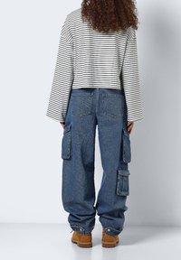 Gestreiftes schwarzes und weißes langärmeliges verkürztes Oberteil, kombiniert mit blauen weit geschnittenen Cargo-Jeans mit Seitentaschen und Bündchen, dazu braune Stiefel.