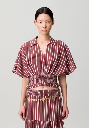 Femme portant un chemisier à rayures verticales bordeaux et blanches avec des manches bouffantes, et une jupe assortie avec une ceinture en pièces d'or, debout devant un fond uni.