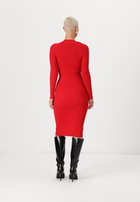 Rød ribbet langermet kjole, tettsittende silhuett, kne-lengde. Kombinert med svarte knelengde stiletthæler. Glatt tekstur, minimal detaljering.