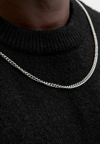 Zilveren ketting die over een zwarte, ribgebreide trui is gedragen. De ketting heeft in elkaar grijpende schakels en een gepolijst afwerking.