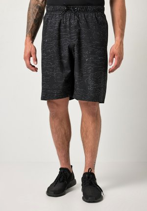 Mand iført sorte atletikshorts med subtilt mønster, sorte sneakers og en sort top, stående på en lys baggrund.