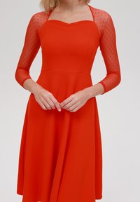 Robe A-line rouge avec de courtes manches transparentes à motif en losanges et un décolleté en coeur. Tissu lisse avec un corps ajusté et une jupe évasée.
