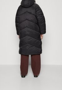 Svart puffjacka med ett chevron-kviltat mönster, med huva och långa ärmar. Matchad med vida bruna byxor och svarta stövlar.