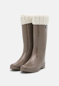 Bottes de pluie brillantes marron avec un revers en tricot crème texturé, semelles plates et design à bout rond. Présente un petit patch de la marque sur le côté.