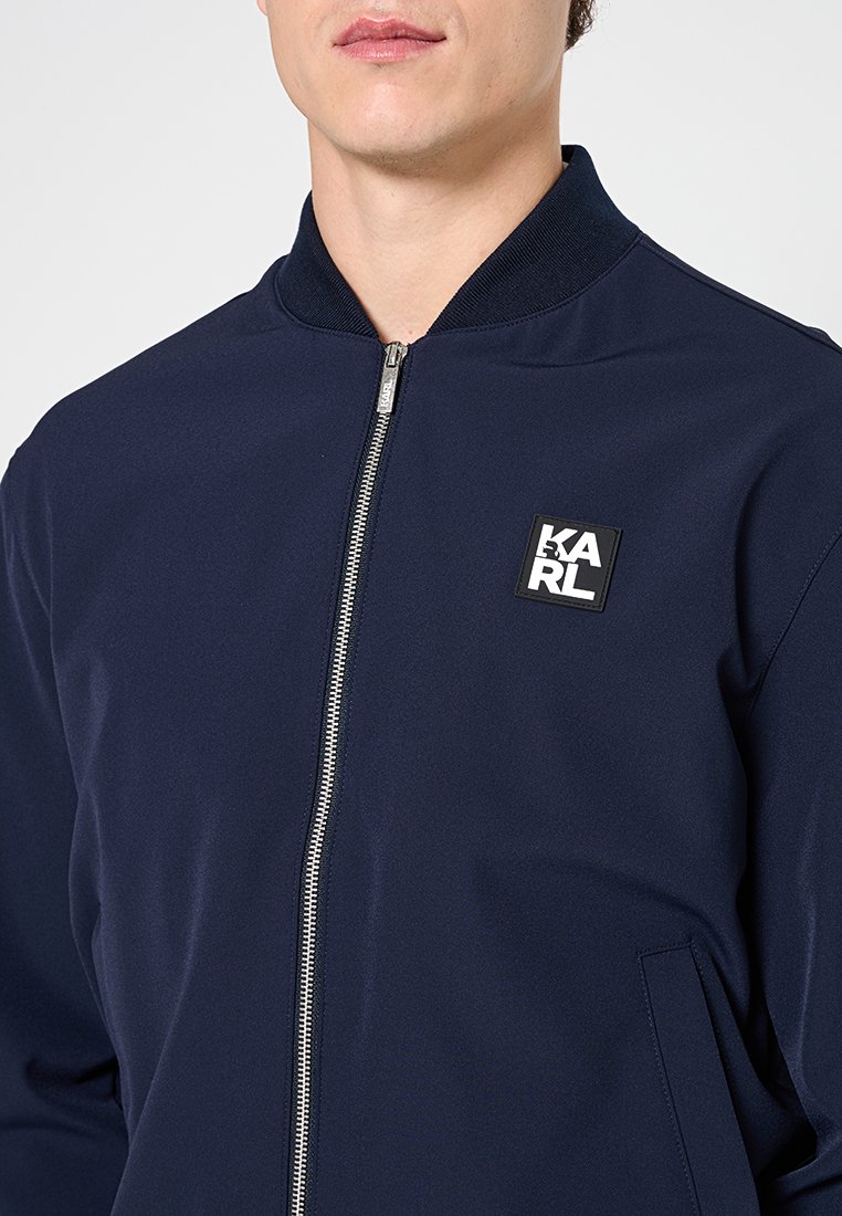 Hombre con chaqueta azul marino con cremallera, cuello acanalado y parche con el logo KARL en blanco y negro en el lado izquierdo del pecho.