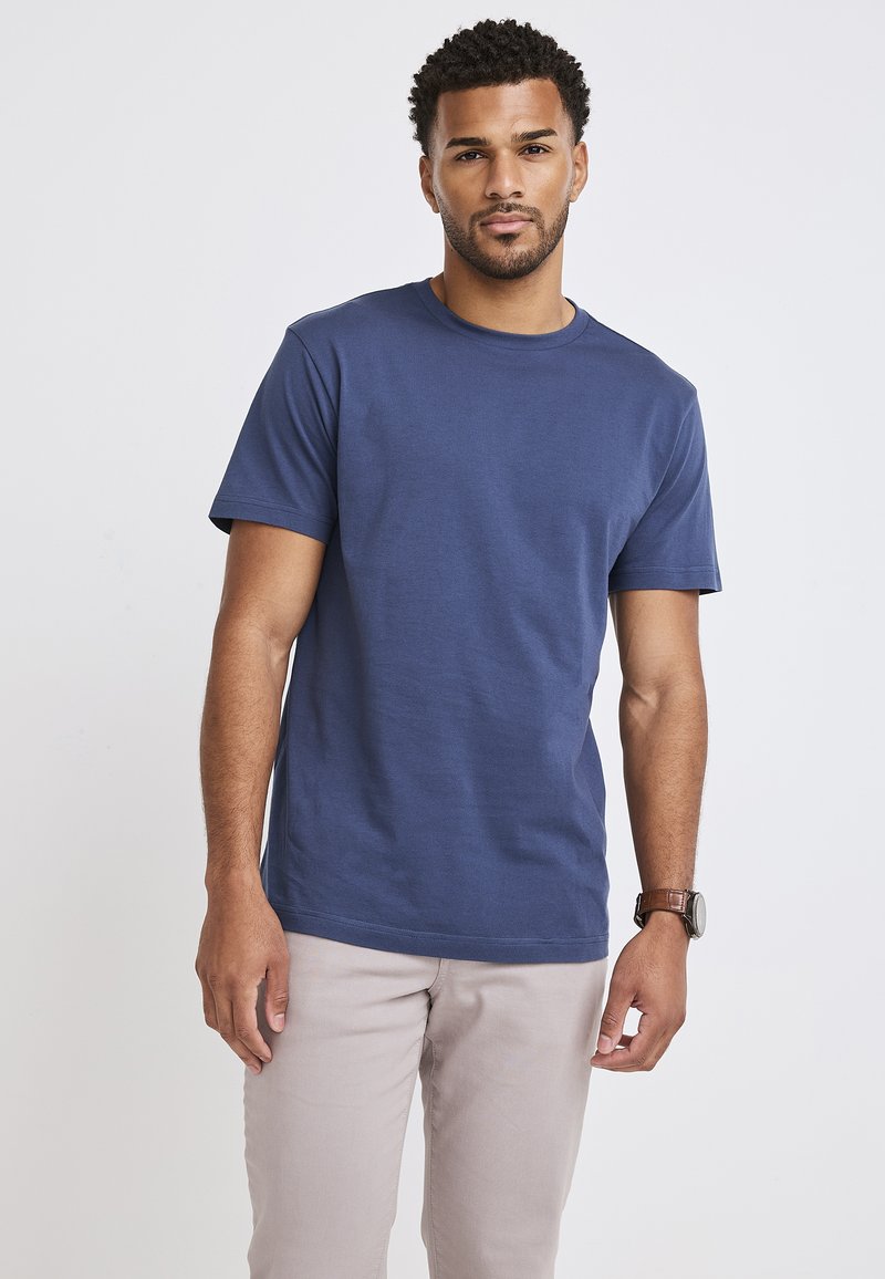 Marinblå bomulls t-shirt med rund hals och korta ärmar; slät textur och rak skärning; använd med ljusbeiga byxor, klocka synlig på handleden.