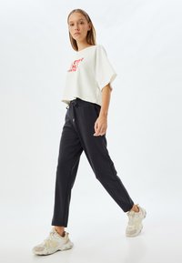 Witte cropped t-shirt met rode tekst, gecombineerd met zwarte joggerpants en lichte sneakers. De stof lijkt zacht en heeft een relaxte pasvorm.