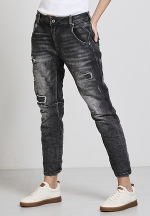 Schwarze Jeans aus Denim mit zerstörten Details, schmalem Schnitt und Fünf-Taschen-Design, kombiniert mit weißen Sneakers mit braunen Akzenten.