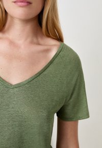 Femme portant un t-shirt vert à col en V à manches courtes, avec la clavicule visible et un teint de peau clair, sur fond uni.