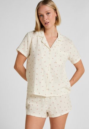 Frau trägt cremefarbenes kurzärmeliges Hemd mit Knöpfen und passende Shorts mit kleinem pinkfarbenem Blumenmuster, steht mit den Händen hinter dem Rücken.