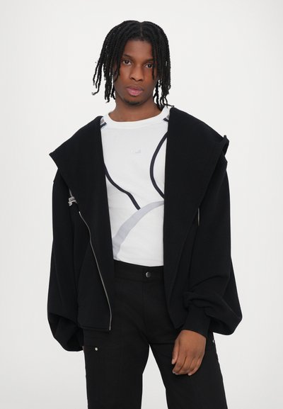 Hanorac supradimensionat, negru, cu închidere cu fermoar, purtat peste un tricou alb cu un design grafic în nuanțe de gri și negru. Pantaloni negri cu buzunare.