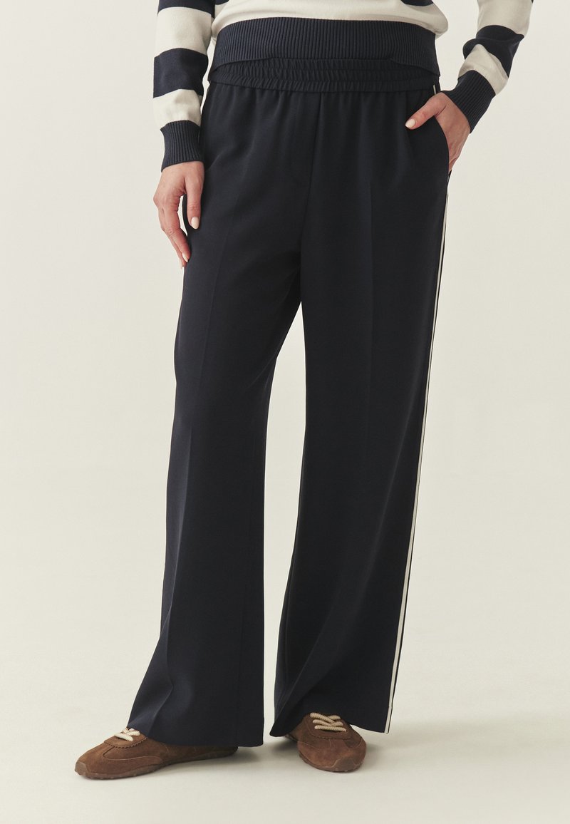 Pantalon bleu marine à coupe large, taille élastique et bandes latérales blanches contrastantes. Le tissu semble doux et léger.