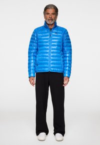 Blå puffjacka med dragkedja och hög krage. Designen inkluderar kviltade paneler, bärs över svarta byxor och vita sneakers.