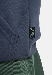 Navyblauwe sweater van textiel met structuur. Voorzien van een zwarte label met witte tekst. Gecombineerd met geribde groene corduroypantalon.