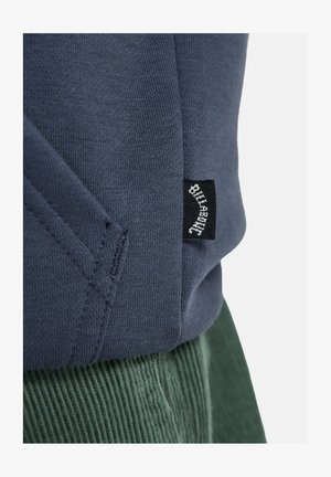 Sötétkék pulóver texturált anyagból. Fekete címke fehér felirattal. Bordó zöld kordbársony nadrággal párosítva.