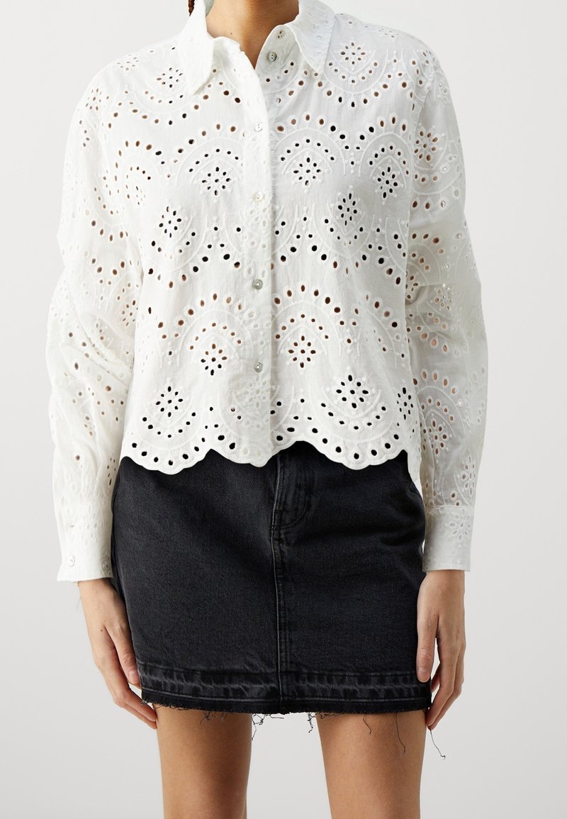 Blouse blanche à manches longues avec œillets et ourlet festonné, associée à une mini-jupe en denim noire effilochée.