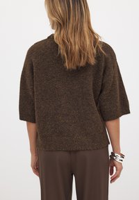 Brauner, strukturierter, locker sitzender Pullover mit kurzen Ärmeln und geripptem Saum, kombiniert mit braunen Hosen und einem silbernen Armschmuck.