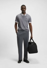 Harmaa polo-paita, jossa on valkoinen kaulus ja hihansuut, yhdistettynä istuviin harmaisiin housuihin. Pitelee mustaa urheilukassia, jalassa mustat kengät.