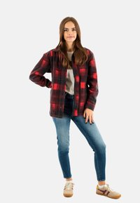 Camisa de franela a cuadros en rojo y negro, diseño de botones, superpuesta sobre una camiseta gris, combinada con jeans azules y zapatillas beige con acentos marrones.