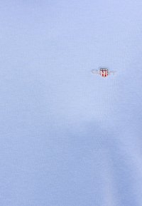 Tissu bleu clair avec un petit logo bouclier brodé présentant des rayures rouges et blanches et des étoiles blanches près du centre à droite.