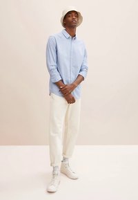 Hellblaues, langärmliges Hemd mit Knöpfen, weiße Hosen mit weitem Bein, weiße Turnschuhe und ein cremefarbener Anglerhut, stehend vor neutralem Hintergrund.