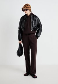 Conjunto desportivo castanho com uma jaqueta bomber de couro preta, um chapéu de balde castanho com padrão, óculos de sol pretos e uma bolsa preta.