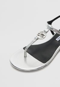 Sølv sandaler med en thong-design, som har en metallisk stropp og sirkulær logo detalj, satt mot en svart såle.