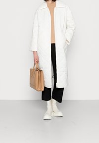 Abrigo largo blanco acolchado con cuello, combinado con una blusa beige, pantalones negros y botas de goma blancas, sosteniendo un bolso marrón claro.