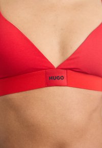 Rød bh med V-hals og bred elastikkant med "HUGO"-logoen i svart. Glatt tekstur, enkelt design.